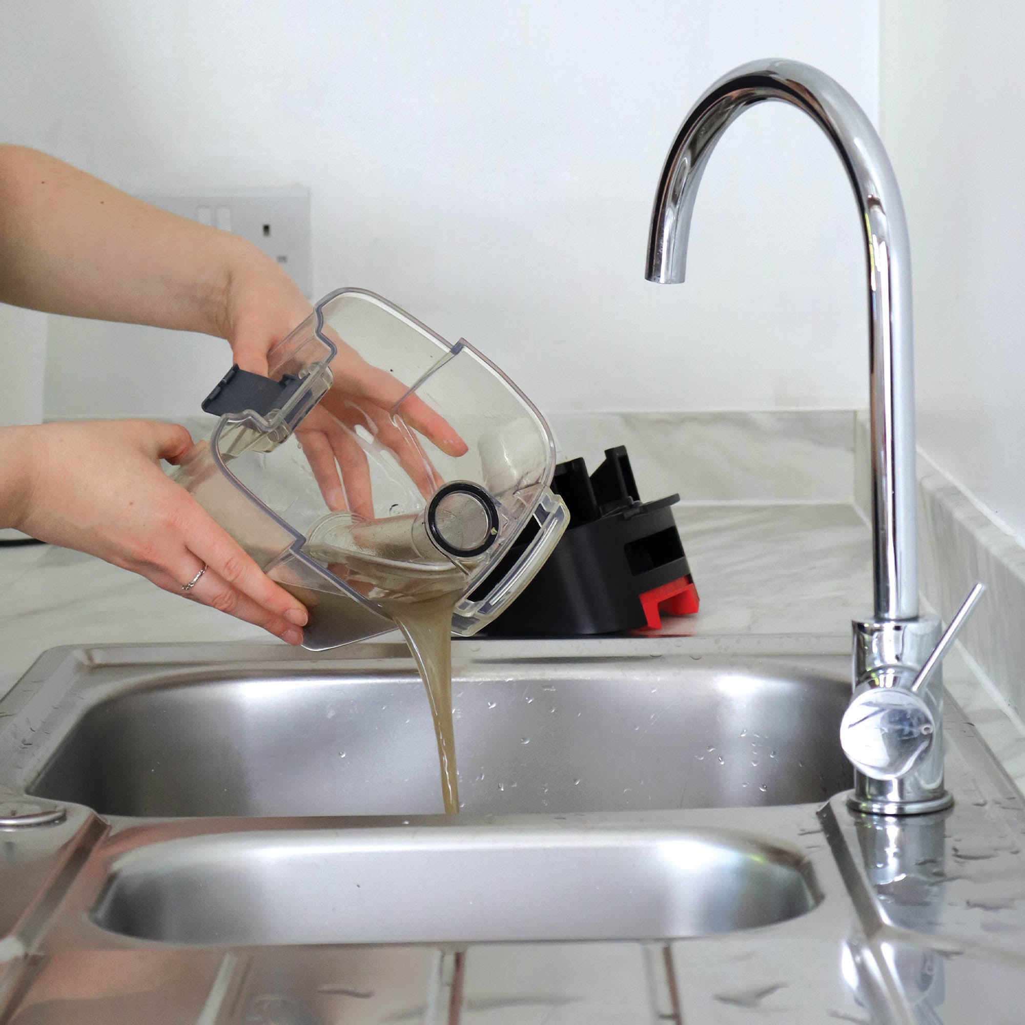 A person empties dirty water from the HYDROC2 Carpet & Upholstery Cleaner into a stainless steel kitchen sink, with the faucet running and water splashing on the countertop.