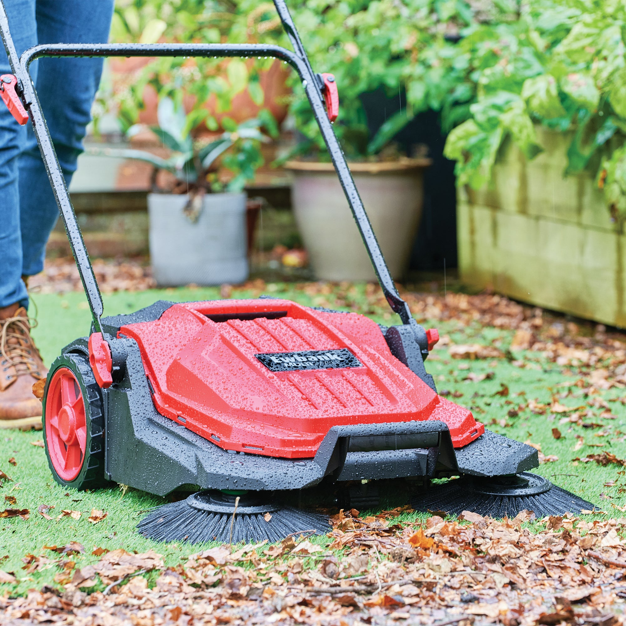A person uses the MULTISWEEP Manual Push Sweeper to clear fallen leaves from a grassy area, with potted plants and garden beds in the background showcasing its effective outdoor cleaning capabilities.