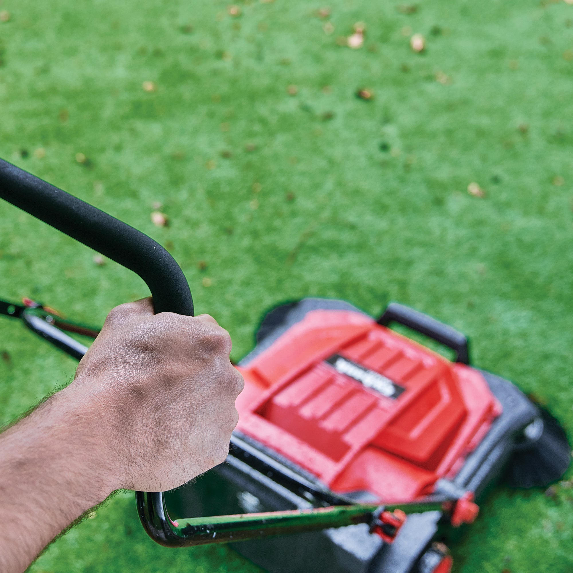 A hand grips the handle of a MULTISWEEP Manual Push Sweeper, moving it across green grass to trim the lawn and handle outdoor cleaning efficiently.