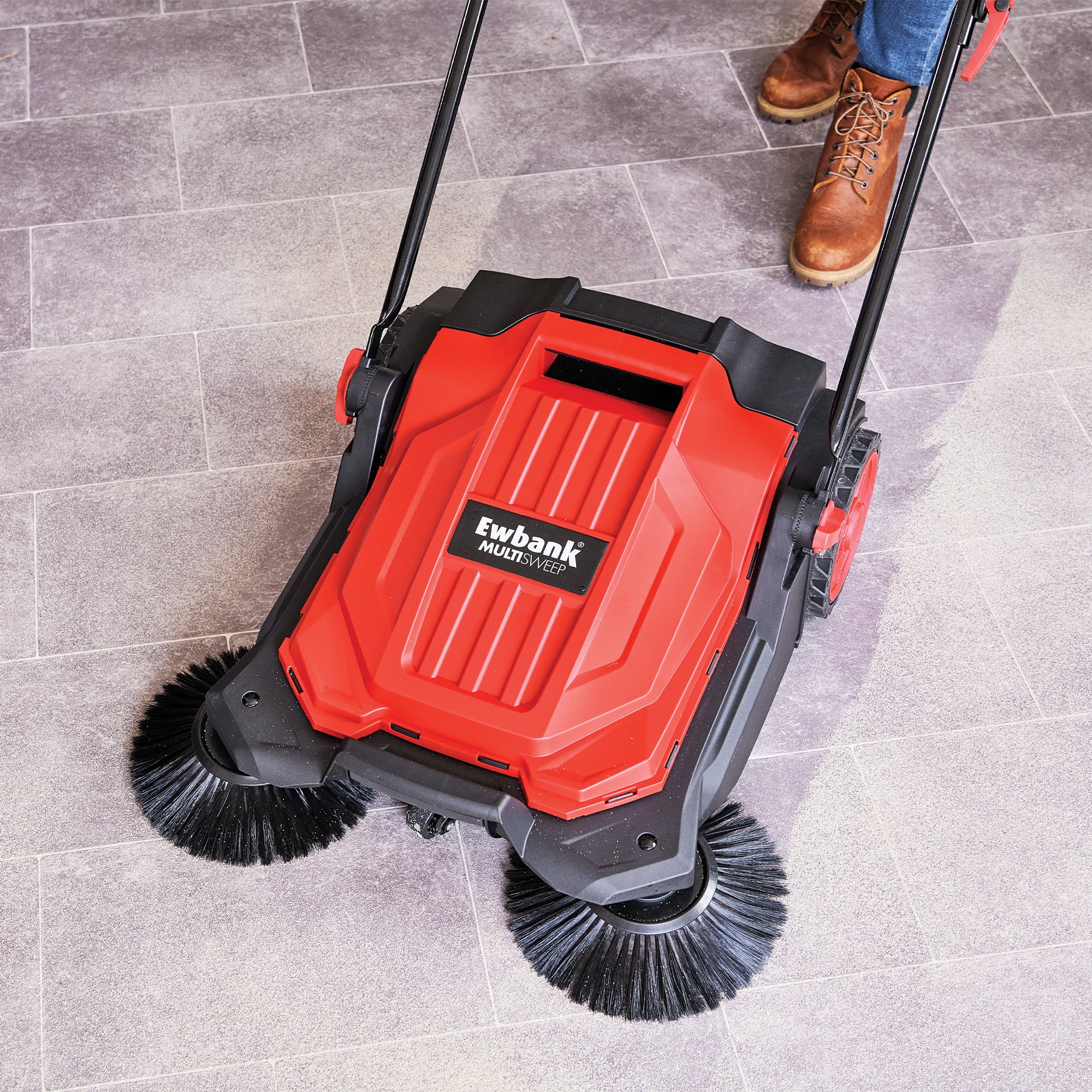 A person in brown boots uses the MULTISWEEP Manual Push Sweeper with two rotating brushes on a gray tiled floor, showcasing its quick and efficient cleaning performance.