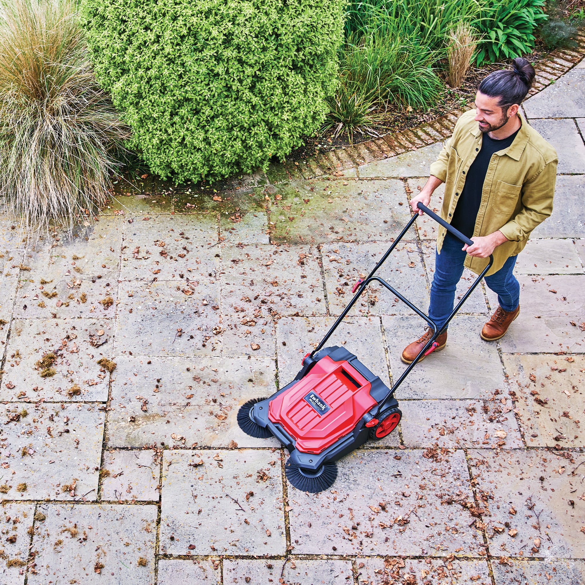 A man with a beard and ponytail uses the MULTISWEEP Manual Push Sweeper, which is red and black, to clean leaves and debris from a stone patio bordered by green shrubs and ornamental grasses.