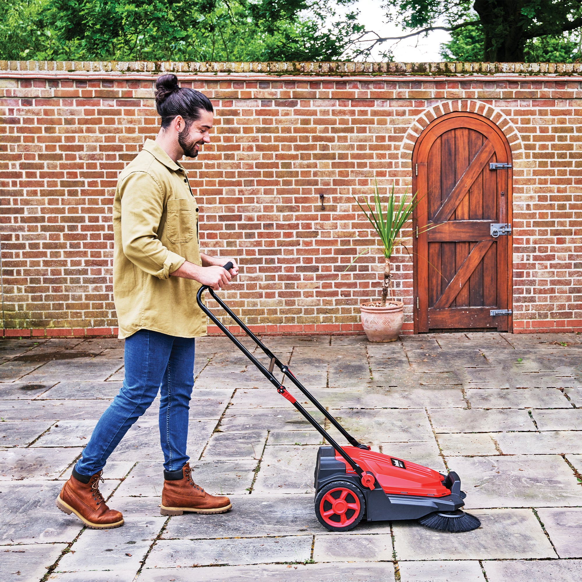 A bearded man with a bun, in a beige shirt, jeans, and boots, uses the MULTISWEEP Manual Push Sweeper to clean a stone patio outside a brick wall with a wooden door and potted plant.