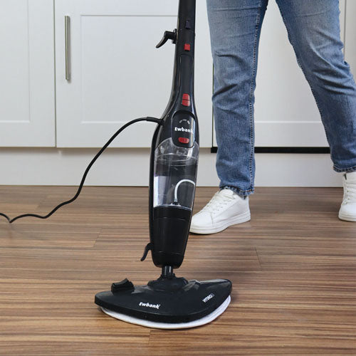 Wearing jeans and white sneakers, a person cleans a wooden kitchen floor with the black Ewbank HYDROS1 5-In-1 Steam Mop; white cabinets are visible in the background.