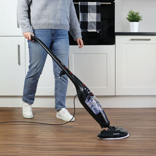 Wearing jeans and a gray sweater, a person cleans a wooden floor in a modern kitchen with white cabinets using the black Ewbank HYDROS1 5-In-1 Steam Mop.
