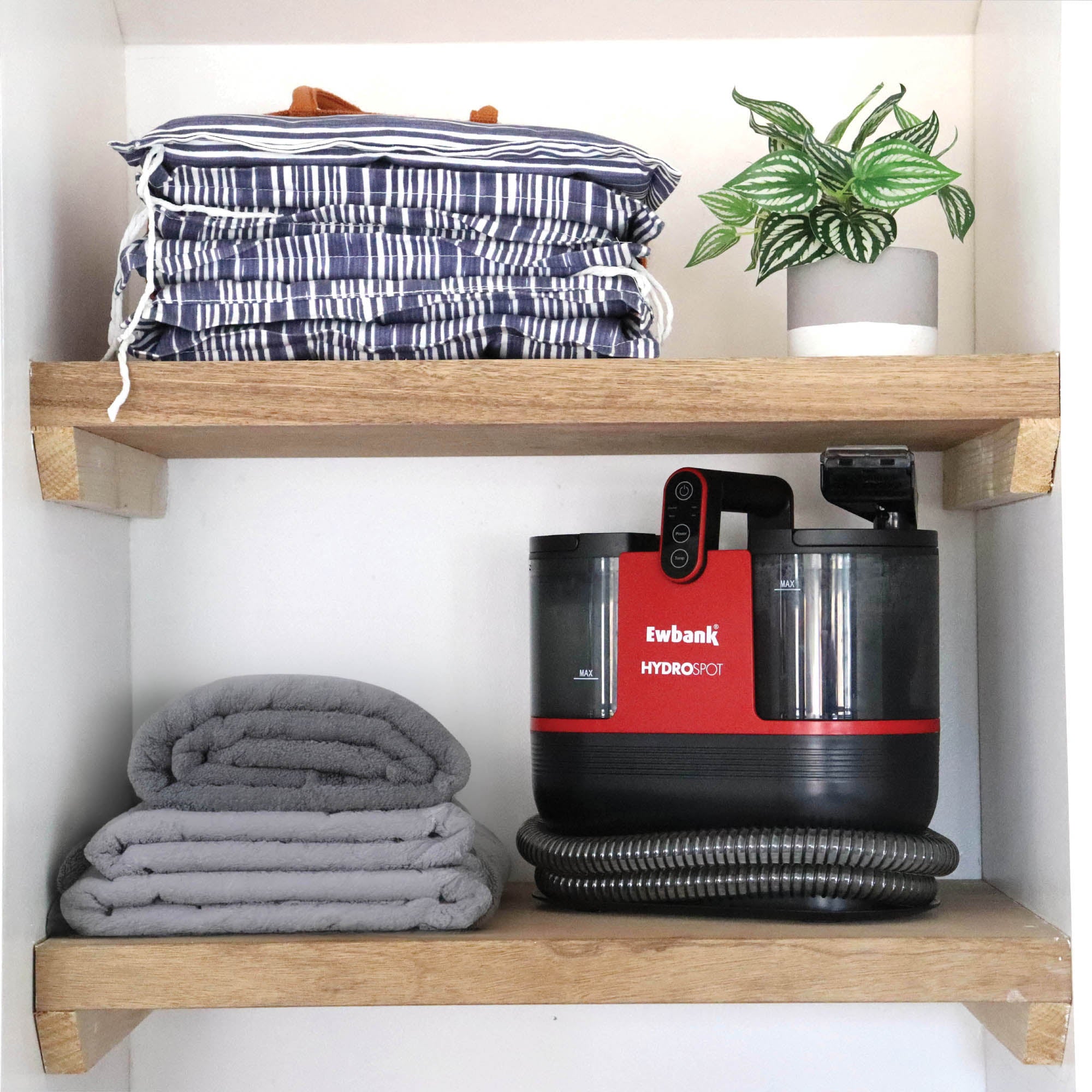 Two wooden shelves display folded striped cushions, a potted plant, gray towels, and the HYDROSPOT Compact Spot Cleaner with hose, all neatly organized within a closet space.