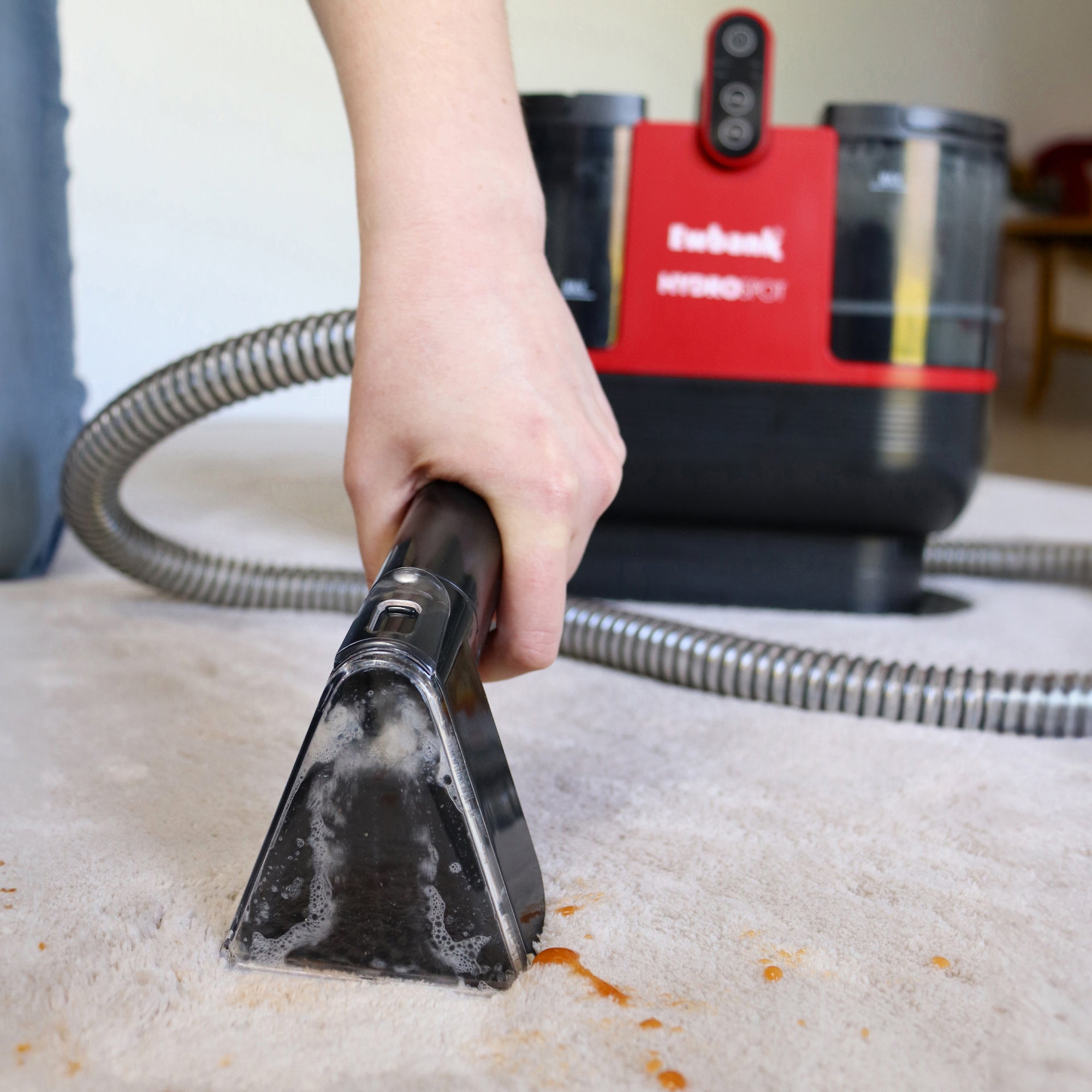 A hand uses the HYDROSPOT Compact Spot Cleaner with a hose to clean stains from a light-colored carpet. The red and black machine is visible in the background, along with soap suds on the carpet.