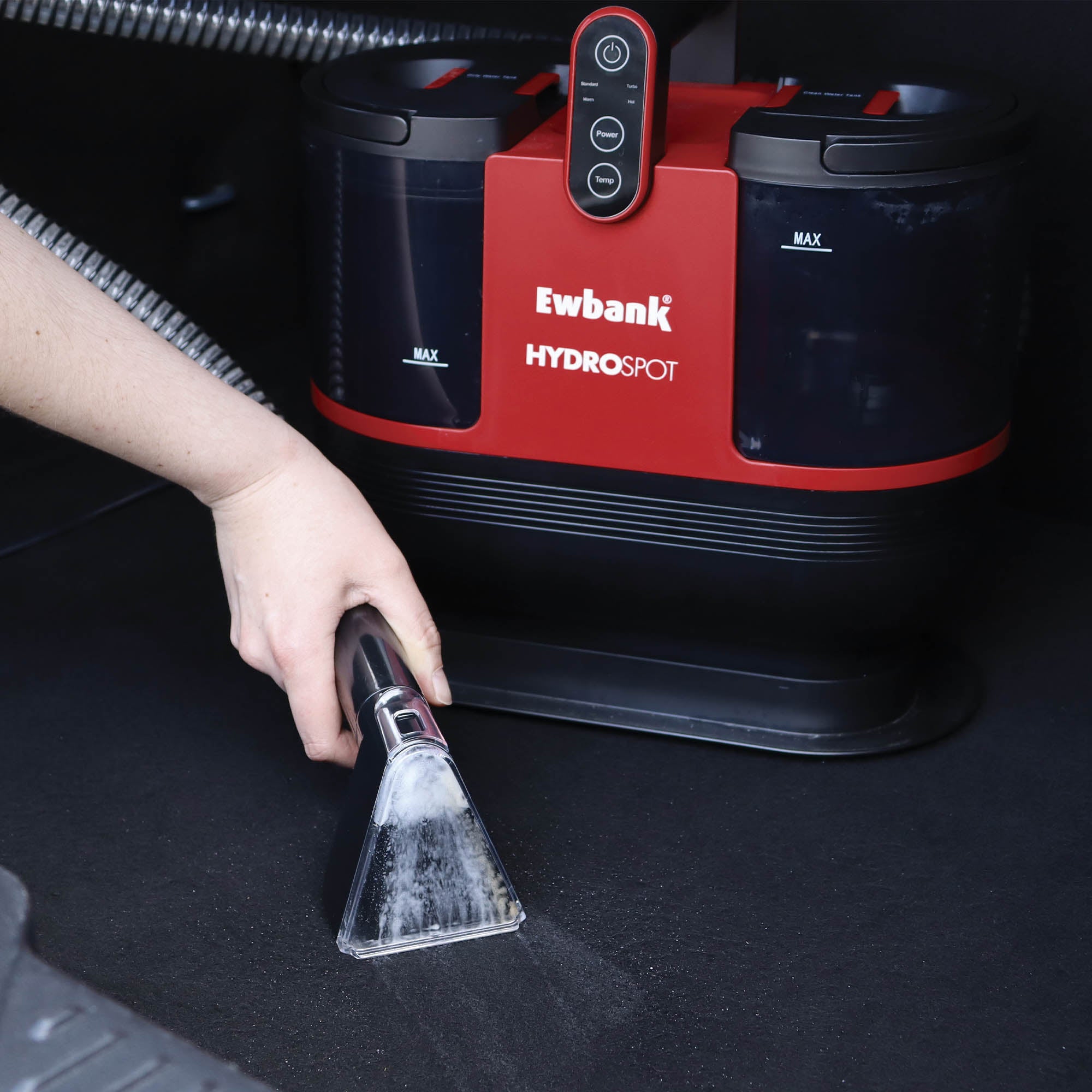 A person uses the HYDROSPOT Compact Spot Cleaner with a handheld vacuum attachment to clean a carpeted surface, effectively removing visible dirt or stains.