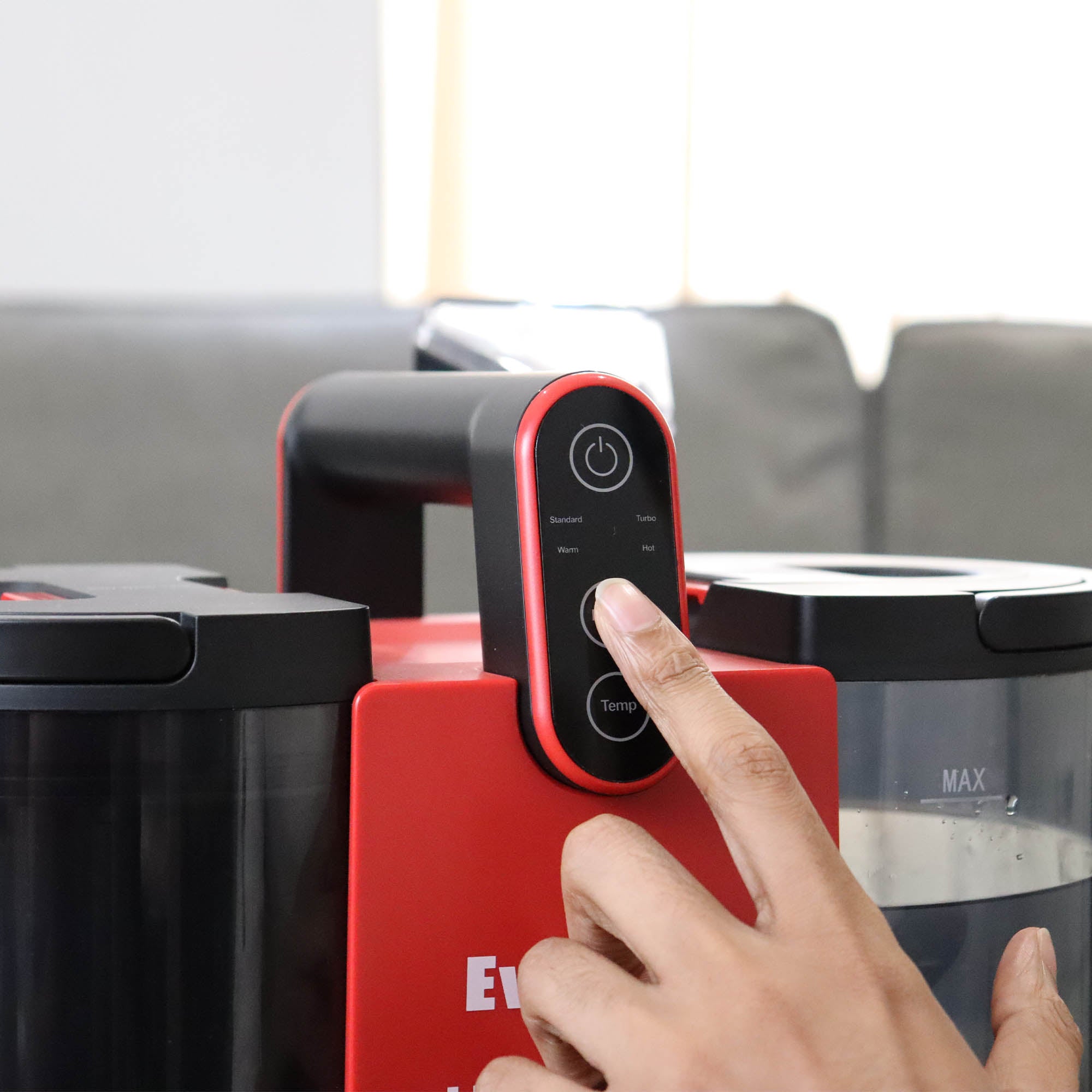 A hand presses a button on the HYDROSPOT Compact Spot Cleaner—red and black with a digital control panel and two containers (one marked “MAX” for water). A couch in the background highlights its upholstery cleaning use.