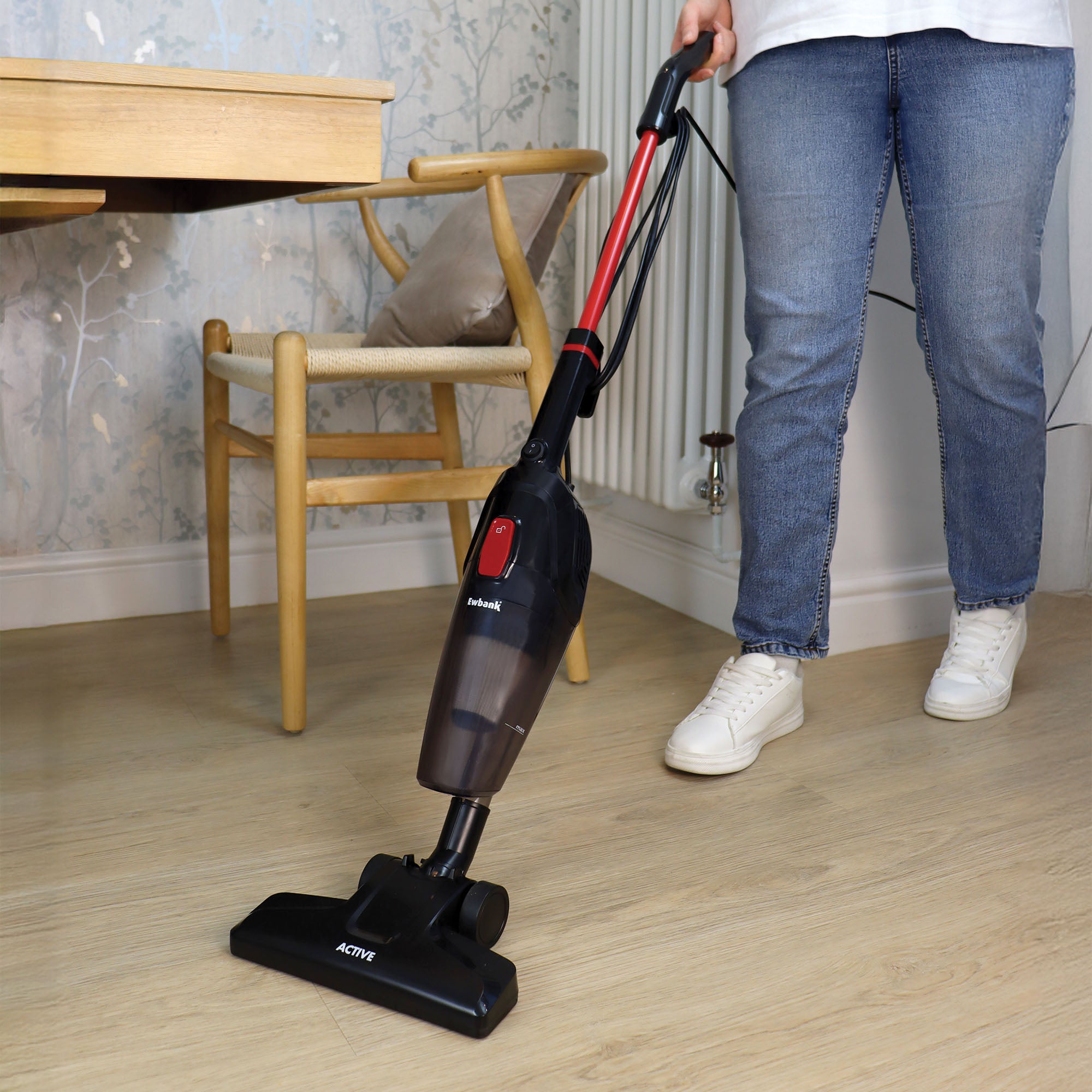 A person in white sneakers and blue jeans vacuums a light wooden floor next to a wooden chair and table using the ACTIVE 2-in-1 Corded Stick Vacuum Cleaner, Red/Black.