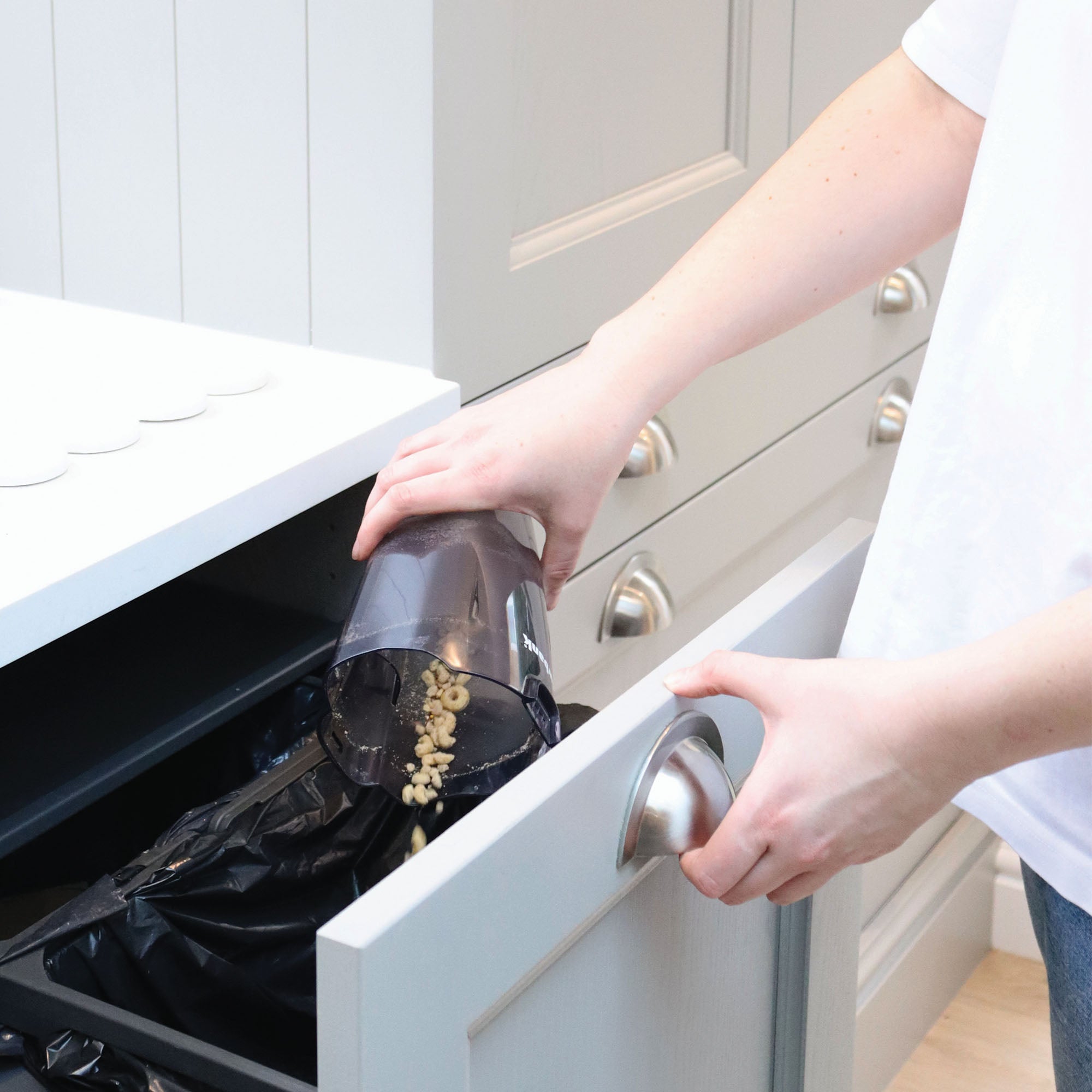 A person empties crumbs from the dust container of the ACTIVE 2-in-1 Corded Stick Vacuum Cleaner, Red/Black, into a trash bin built into a light gray kitchen cabinet.