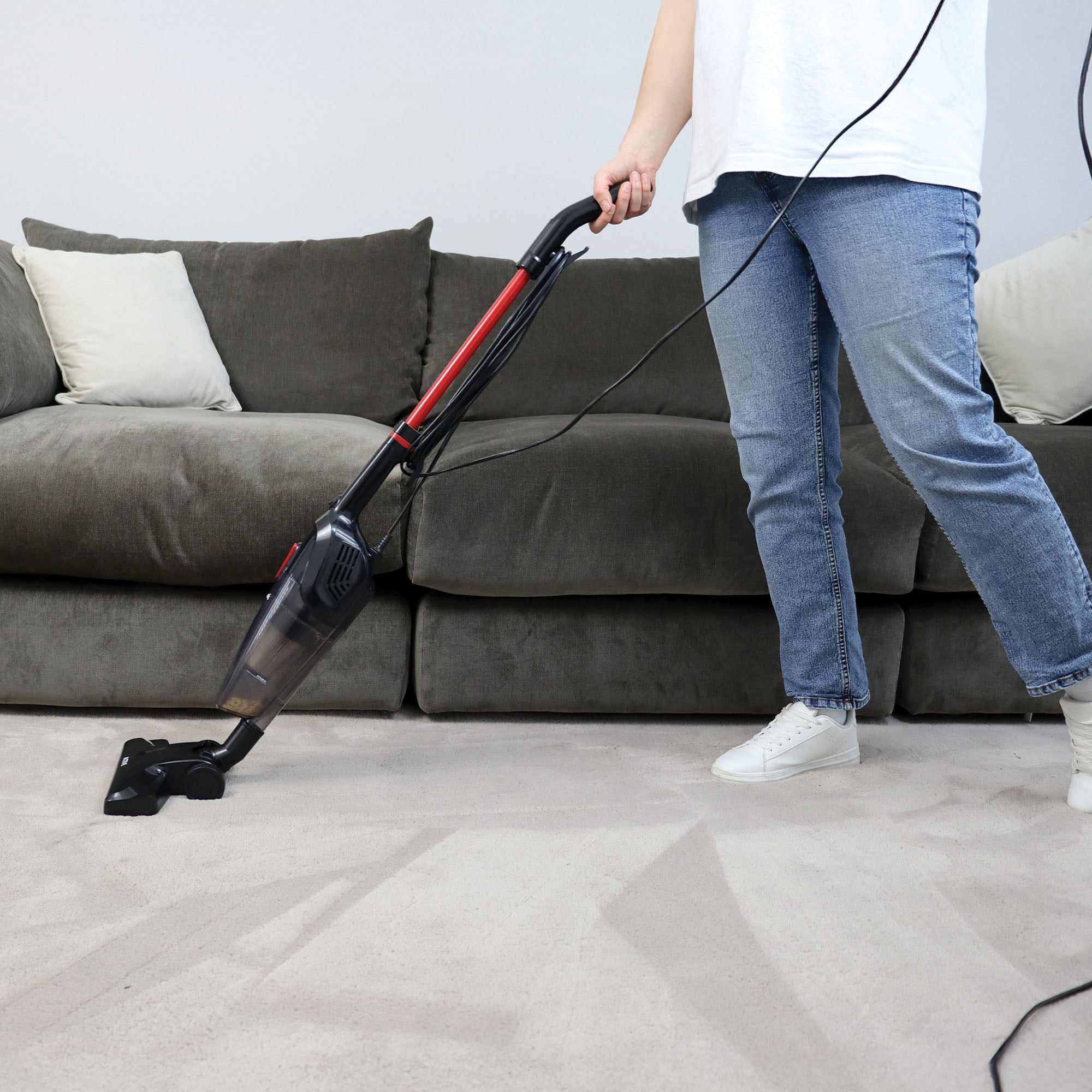 A person in jeans and white sneakers uses the ACTIVE 2-in-1 Corded Stick Vacuum Cleaner, Red/Black, to clean a light-colored living room carpet, with a gray sofa and beige cushions visible in the background.