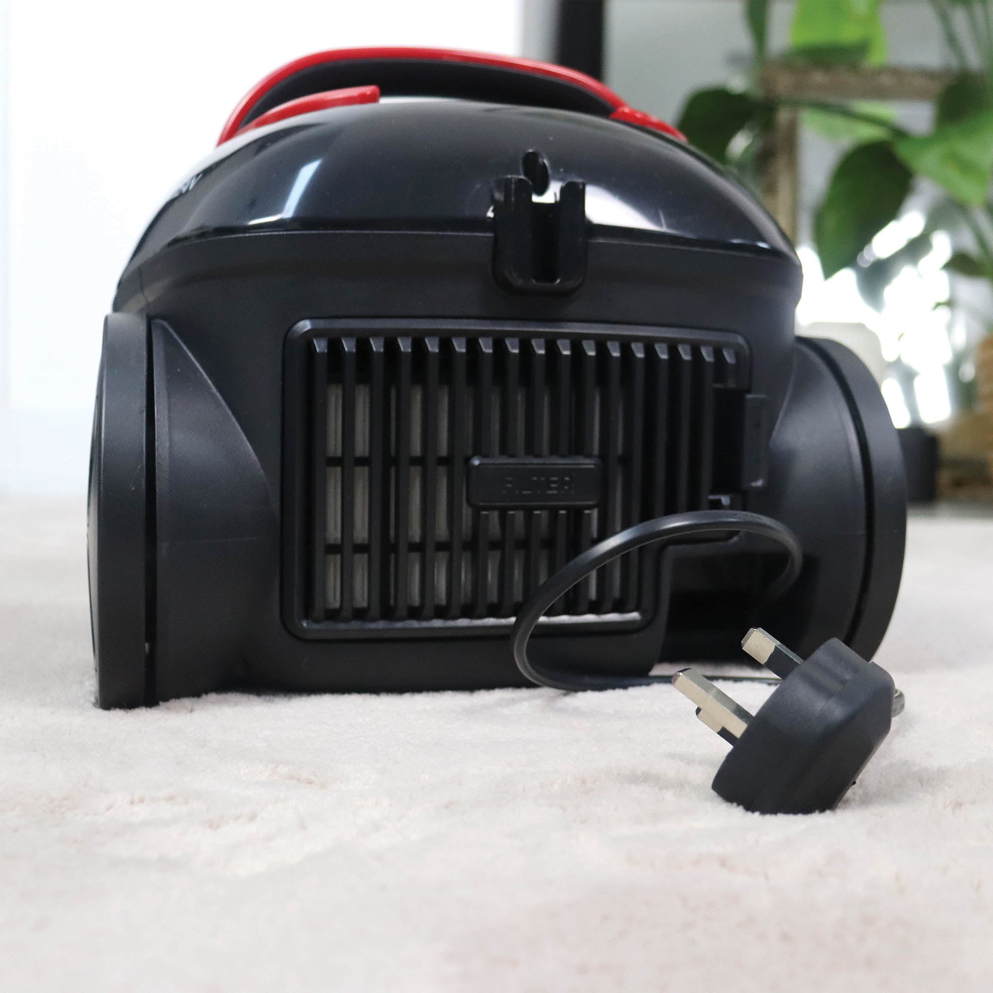 The Ewbank COMPACTCLEAN Bagged Vacuum Cleaner in black rests on a light carpet, its power cord and two-prong plug visible in front. Blurred green indoor plants appear in the background.