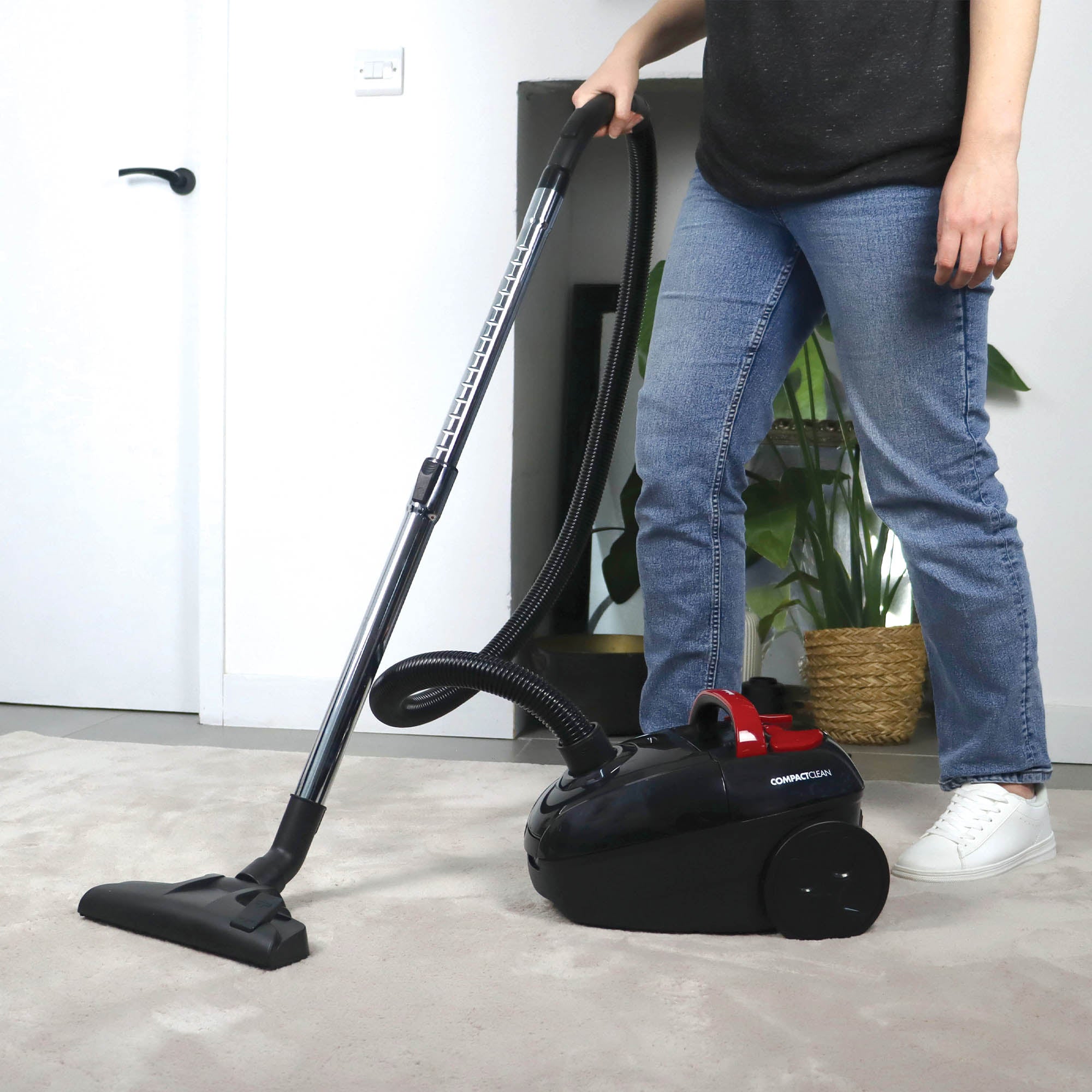 A person in jeans and white shoes is vacuuming a carpet with the Ewbank COMPACTCLEAN Bagged Vacuum Cleaner, which features a HEPA filter, in a bright modern room with a potted plant visible in the background.