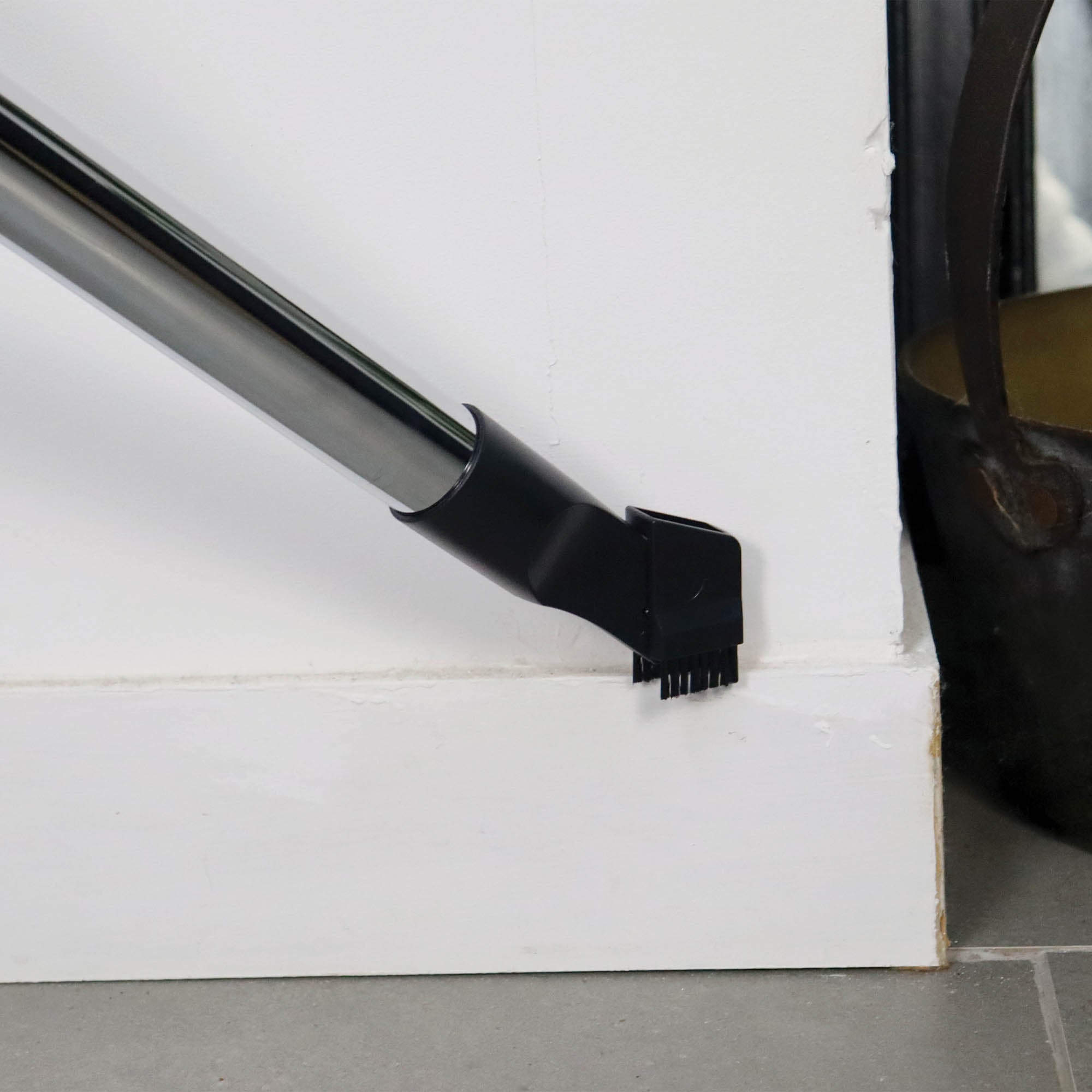 The Ewbank COMPACTCLEAN Bagged Vacuum Cleaner, equipped with a narrow brush attachment, is cleaning along the edge of a white baseboard near a corner wall.