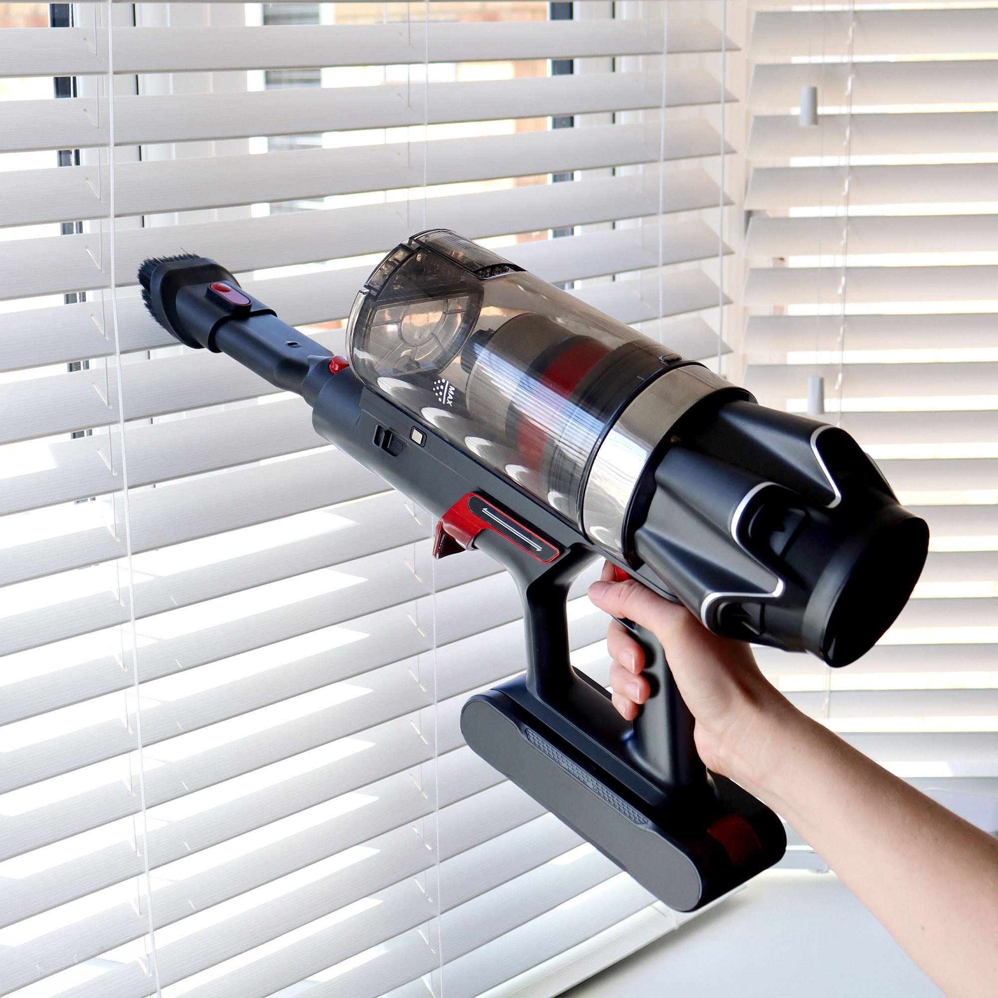 A person holding the AIRSTORM AUTO Empty Cordless Stick Vacuum Cleaner with its reusable dust bag near white window blinds, preparing to clean them.