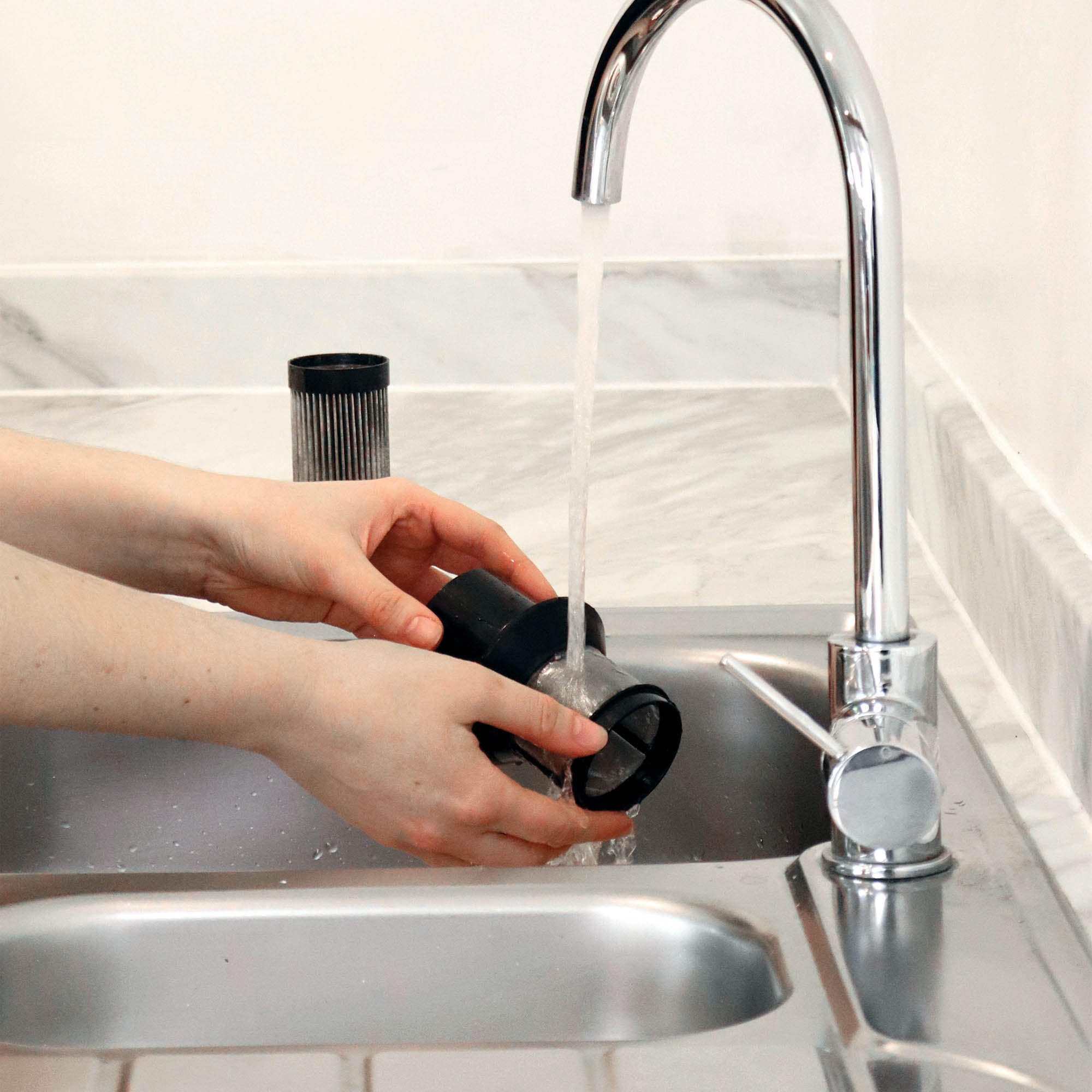 A person rinses a washable HEPA filter from the AIRBLITZ 2-in-1 Cordless Stick Vacuum Cleaner under a kitchen tap, while another filter part stands upright on the countertop nearby.