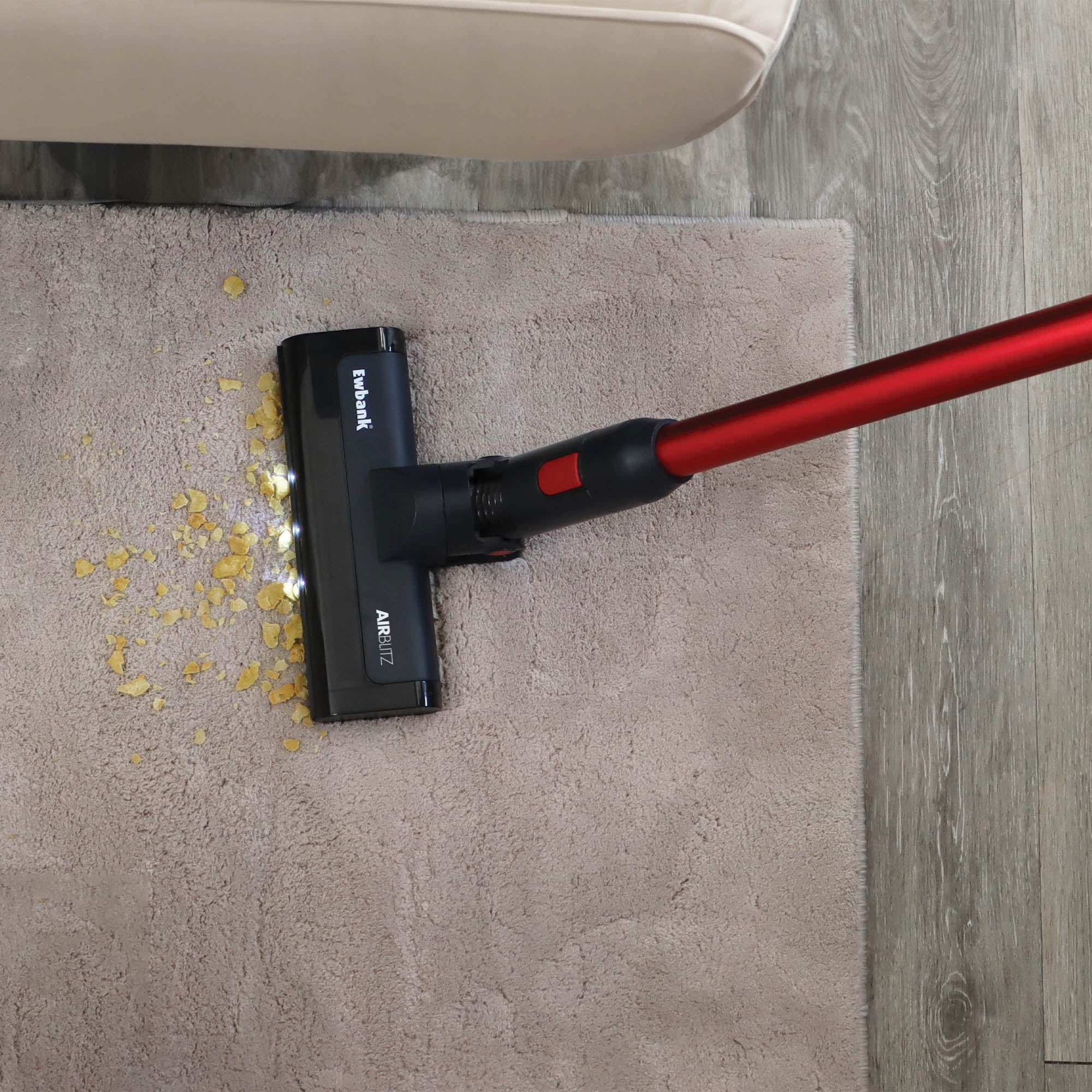 The AIRBLITZ 2-in-1 Cordless Stick Vacuum Cleaner with a red handle cleans up crumbs from a light brown rug near a beige sofa on a wood floor.
