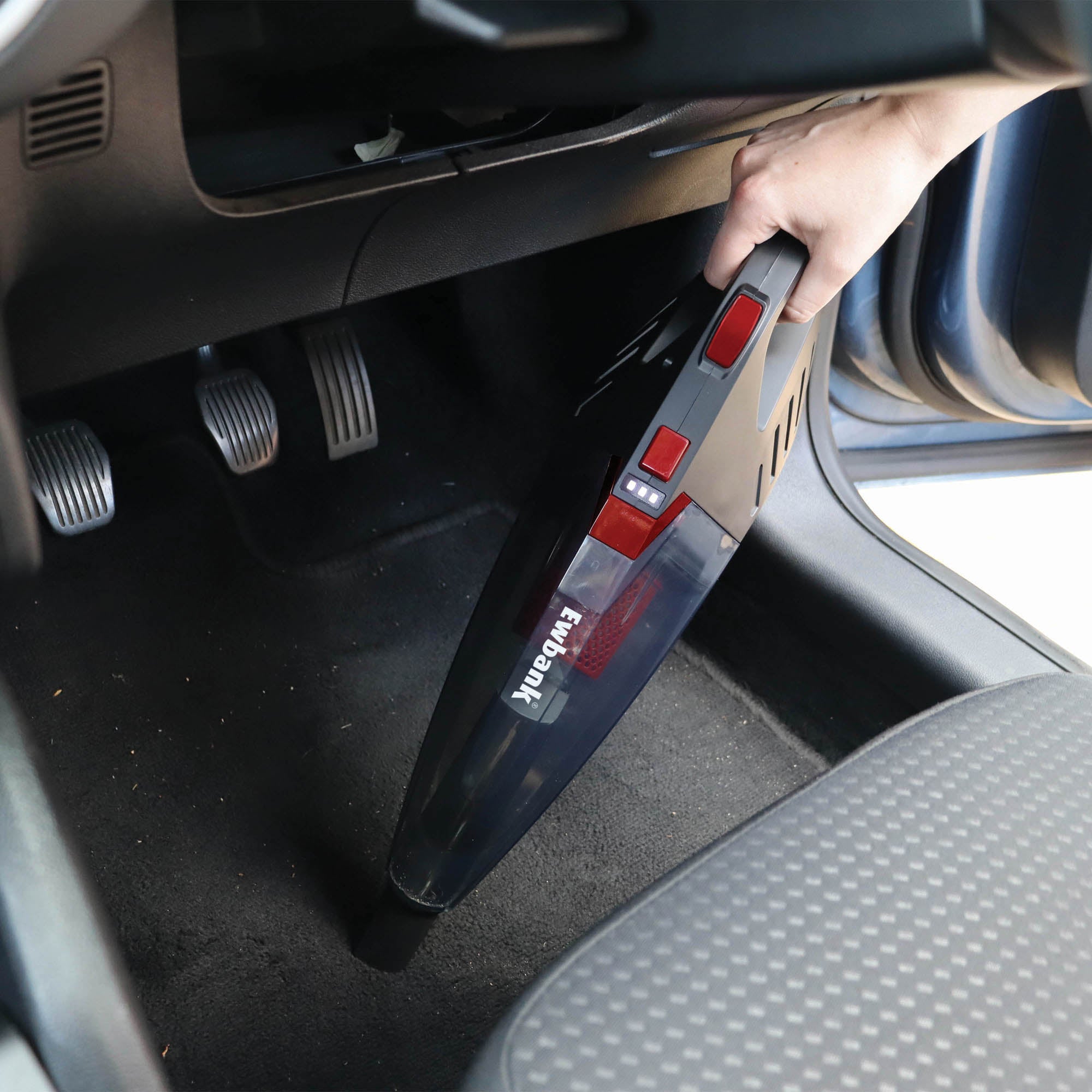 A person uses the ACTIVE+ 2-in-1 Cordless Stick Vacuum Cleaner to clean the floor mat area under a car’s dashboard, near the pedals and front passenger seat.