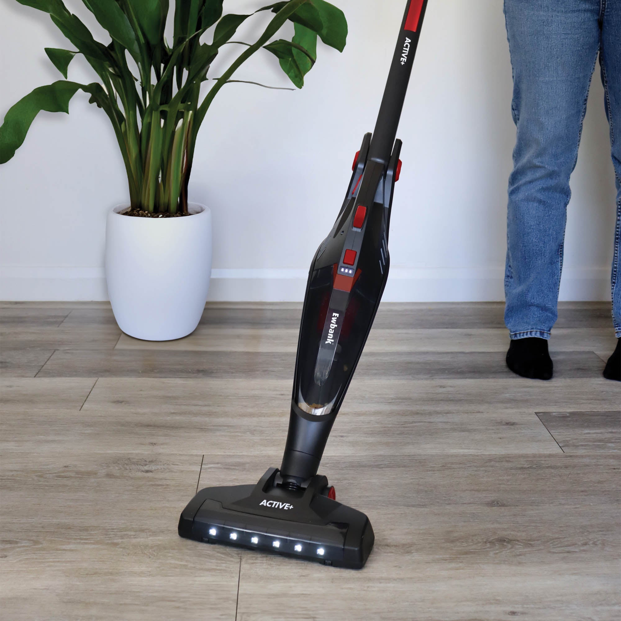 A person in jeans uses the ACTIVE+ 2-in-1 Cordless Stick Vacuum Cleaner with a HEPA filter on a light wooden floor near a green plant in a white pot.