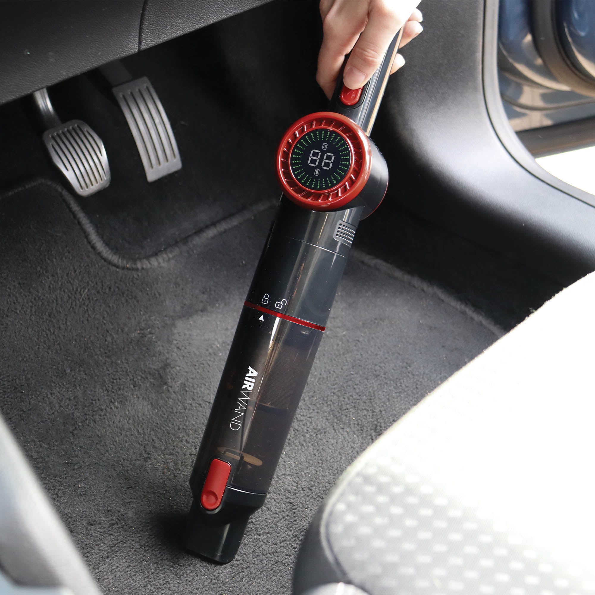 A hand uses the AIRWAND Cordless Handheld Vacuum Cleaner with a brushless DC motor to clean a car floor mat near the pedals. The vacuum, black and red in color, displays a digital number on its top.