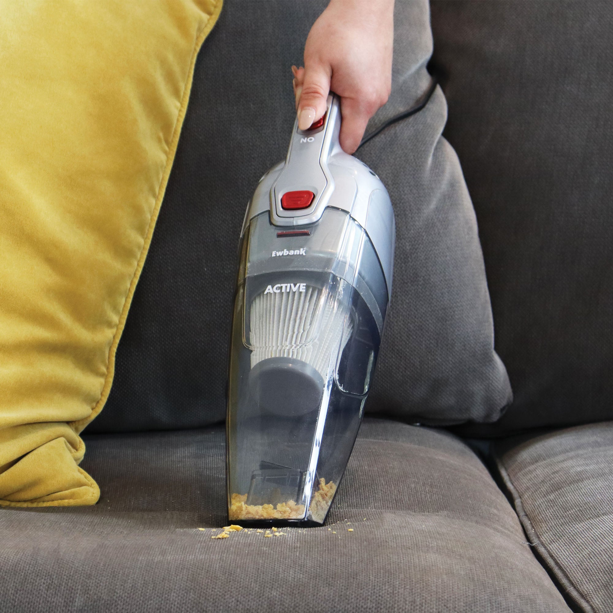 A hand uses the ACTIVE 2-in-1 Corded Stick Vacuum Cleaner to pick up crumbs from a gray sofa near a yellow pillow—ideal for cleaning all floor types.