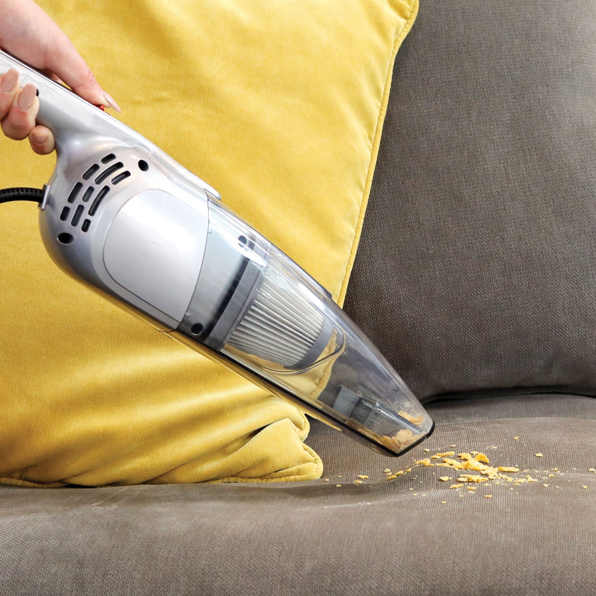 A person uses the ACTIVE 2-in-1 Corded Stick Vacuum Cleaner to remove crumbs from a gray couch with a yellow cushion.